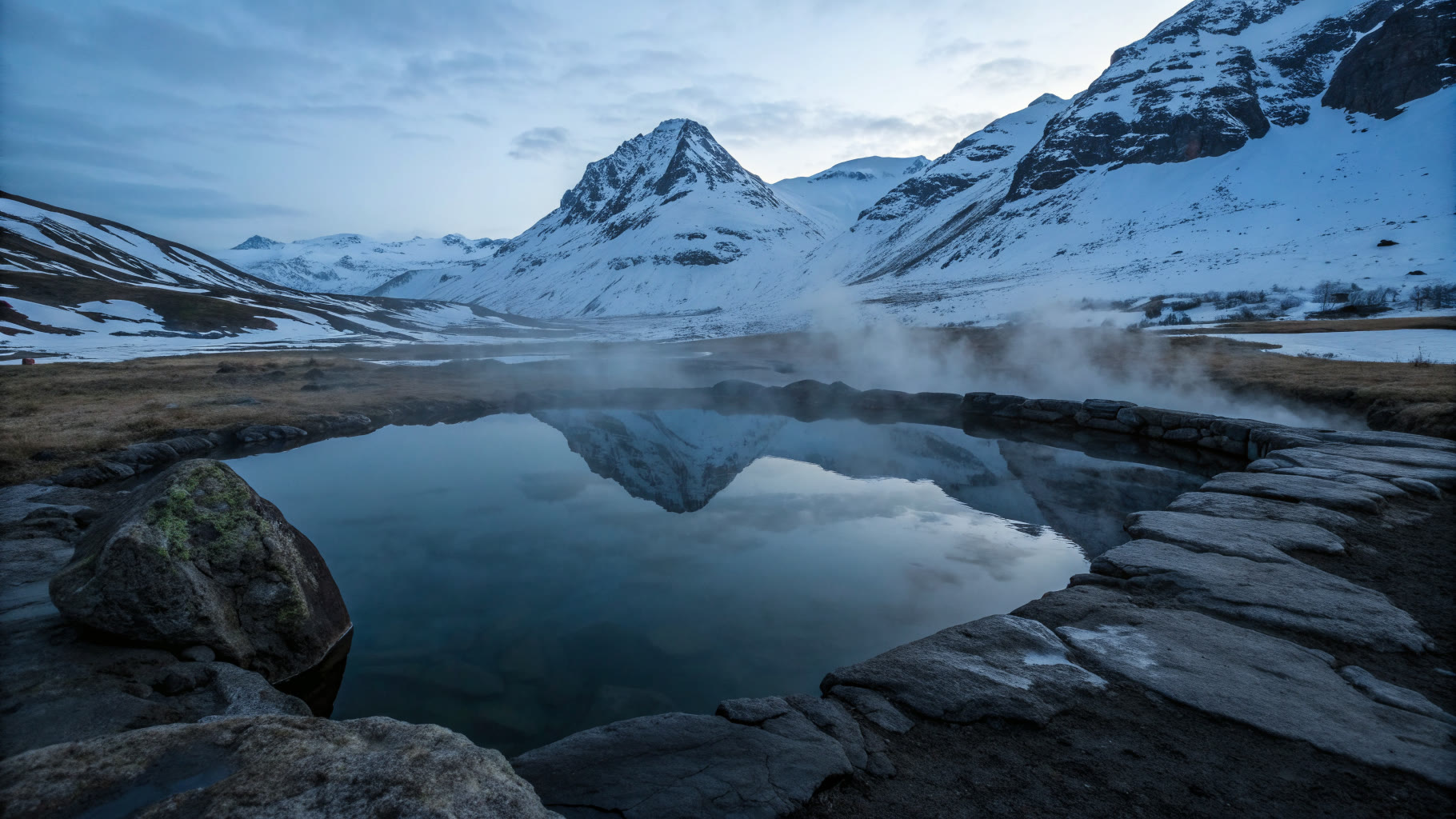 Vasca termale all'aperto che fuma nell'ora blu, con le cime innevate riflesse nell'acqua ferma